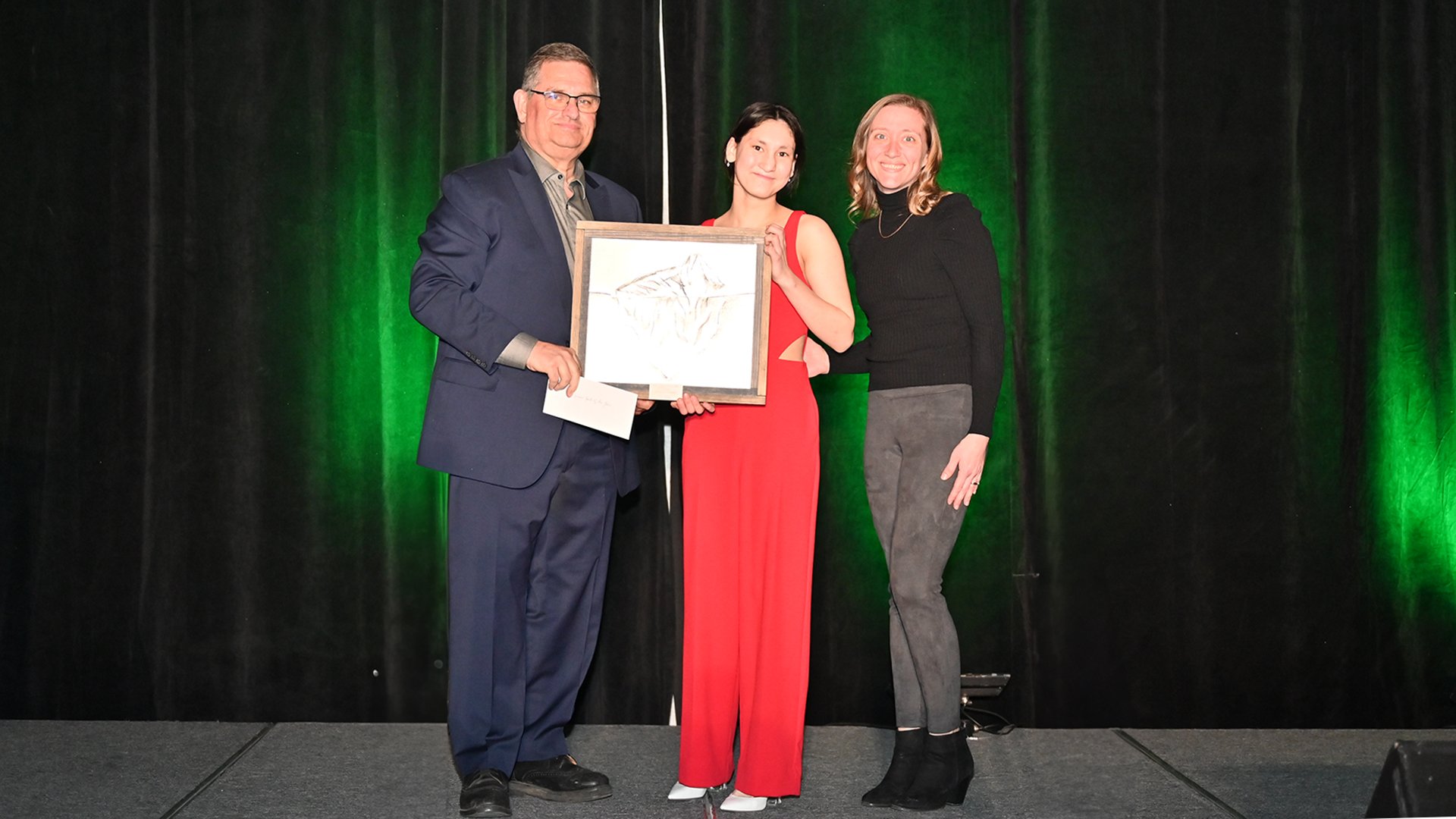 A man left with two women on stage, woman in the middle holds an award