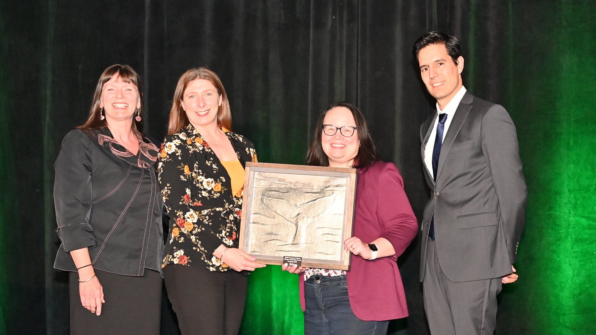 Three woman and a man stand on stage, two women in middle hold an award