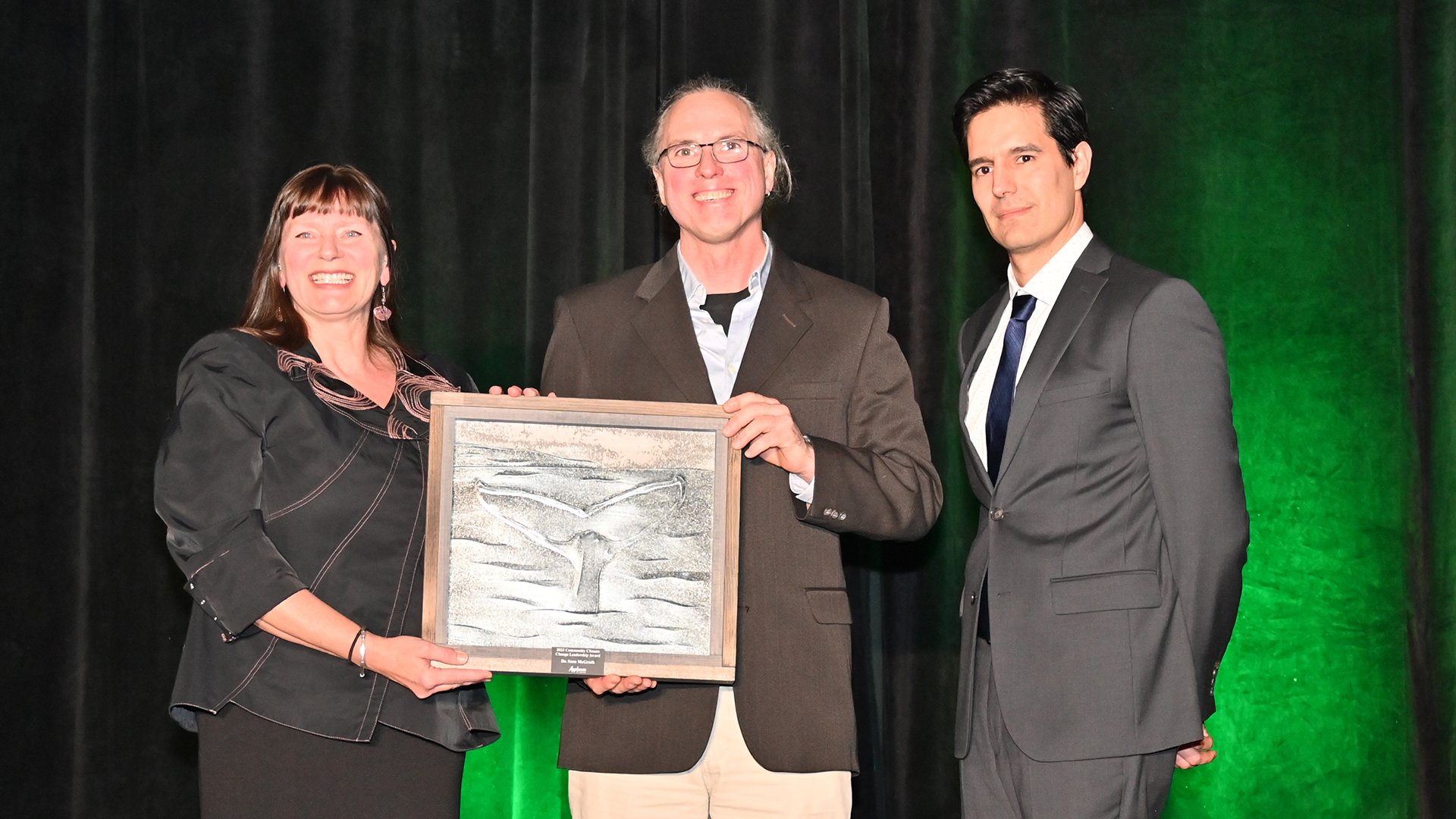 Two men and a woman stand on stage. A man in the middle holds and award