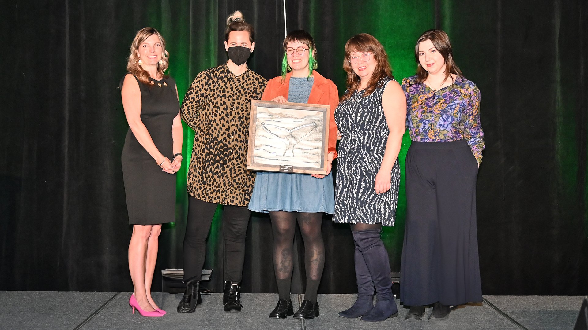 A group of five women stand on stage, the woman in the middle holds an award