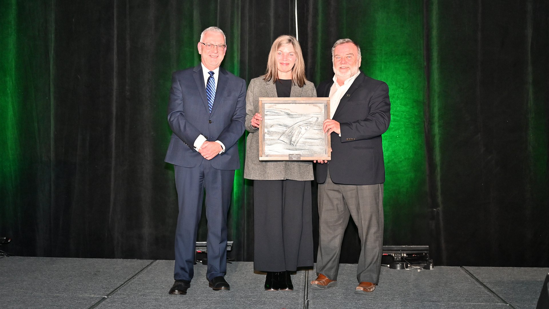 Two men and a woman stand on stage, woman in the middle holds an award