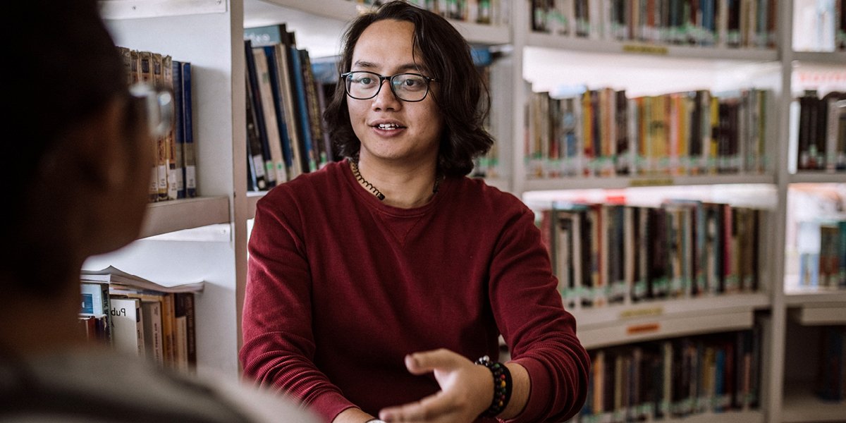 Person sitting in front of book shelves talking to another person. 