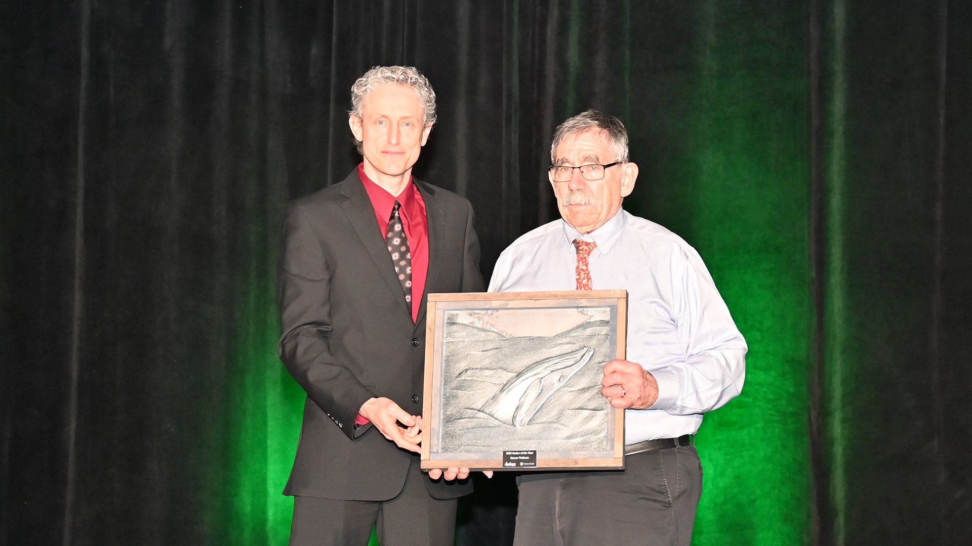 Two men stand on stage, one at right holds an award