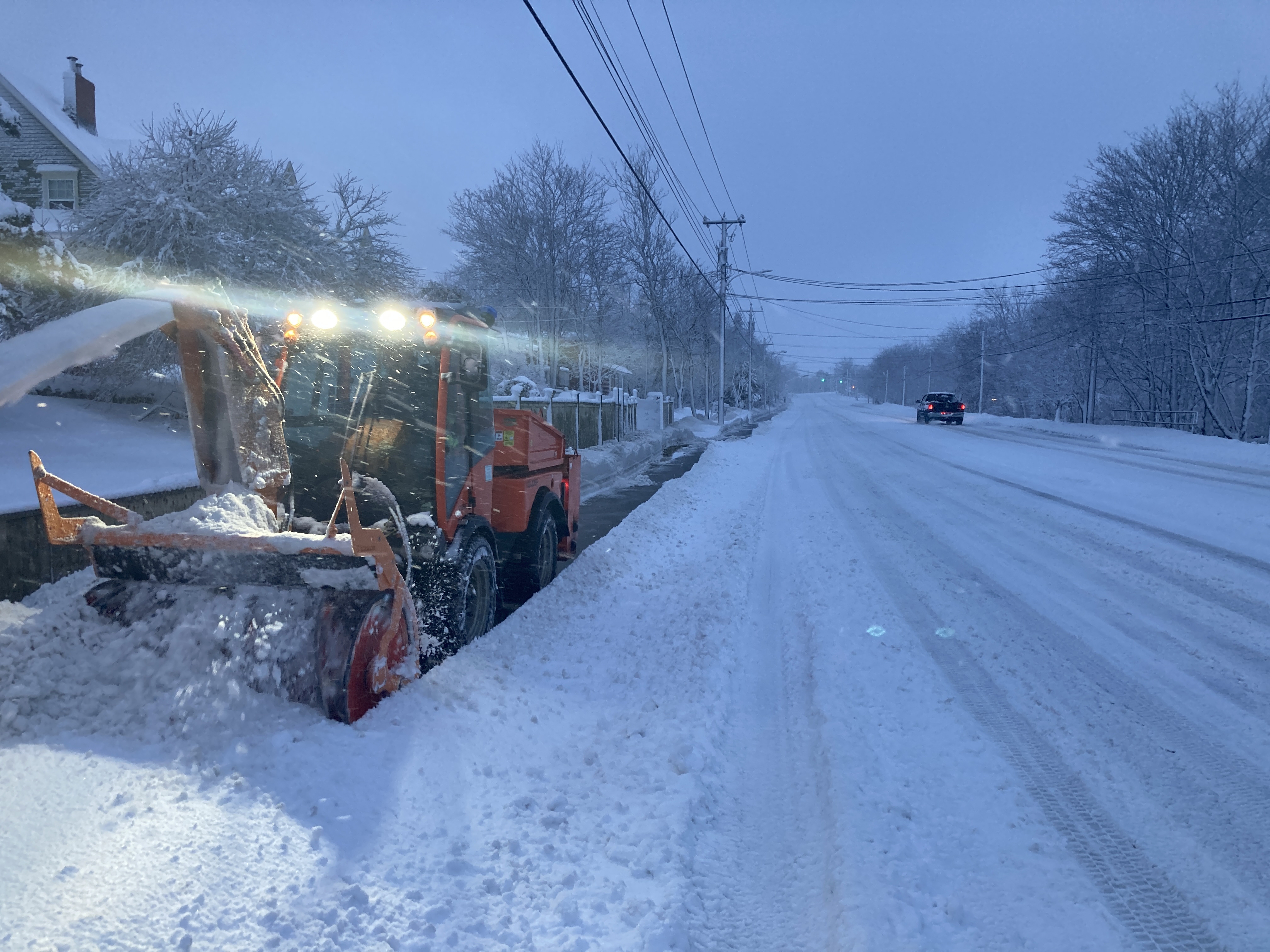 A sidewalk snow plow on the left of the image, and a snow covered road on the right.