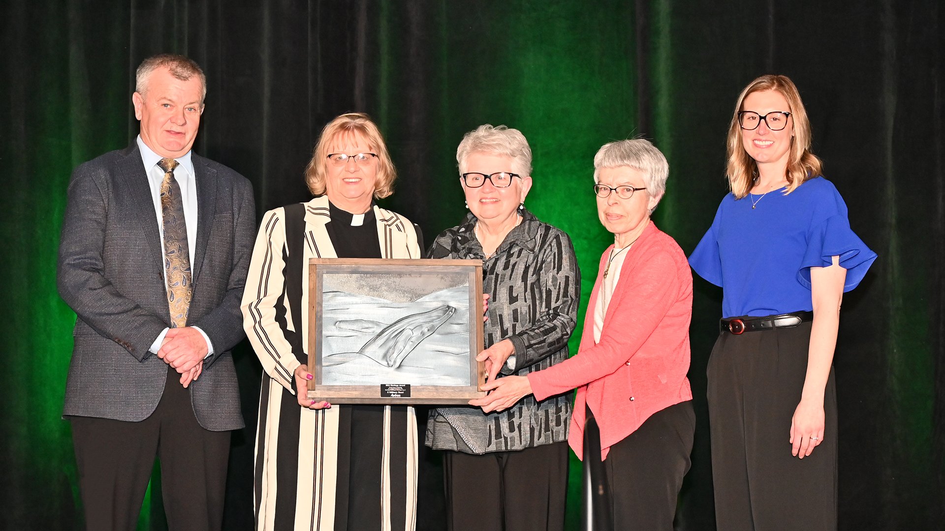 Four women and a man stand on stage. Three women in the middle hold an award.