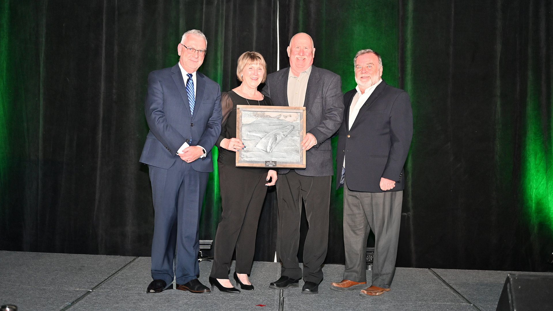 Three men and a woman stand on stage. Man and woman in the middle hold an award