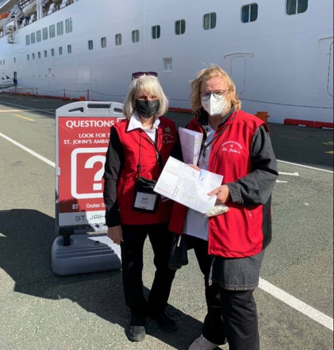 two individuals wearing masks in red vests smile in front of a cruise ship