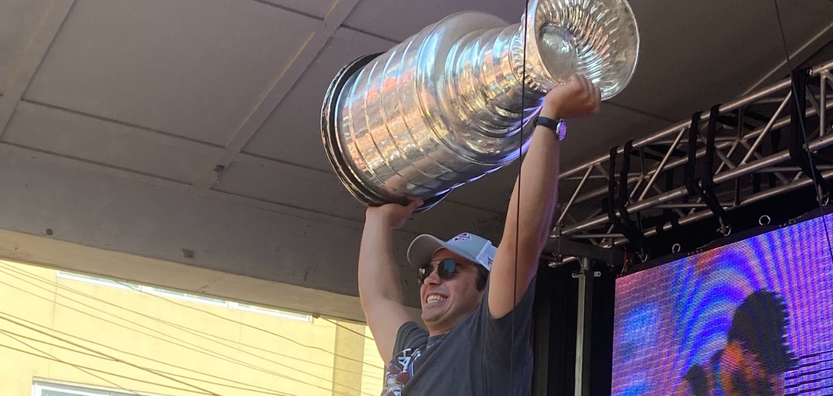 Alex Newhook, smiling, hoists Stanely Cup above his head on George Street