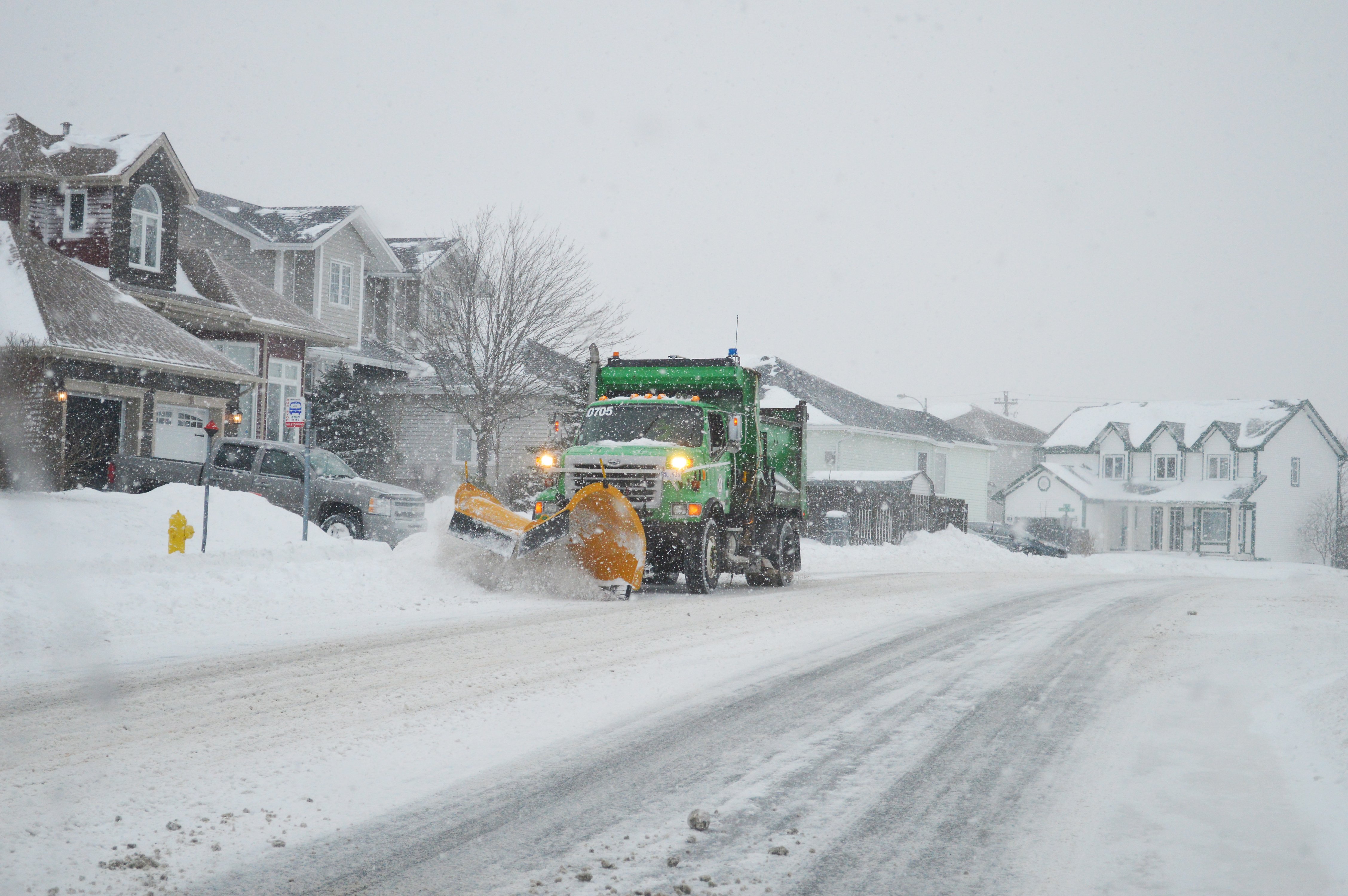 A snow covered city street, with homes in the background. A green snow plow with a yellow blade is on the left of the image.