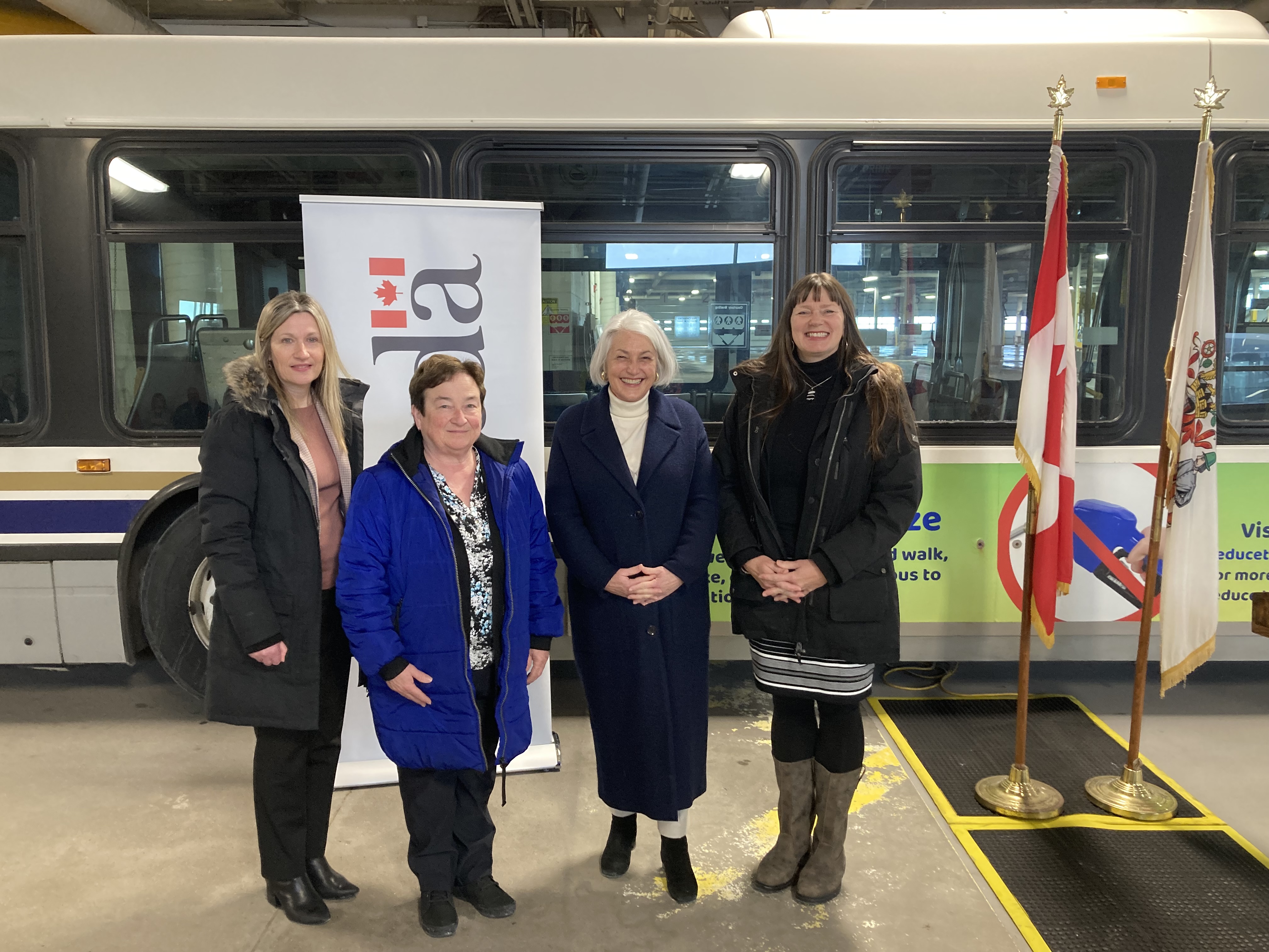 Four people standing together in front of a city bus, there are two flags in the background.