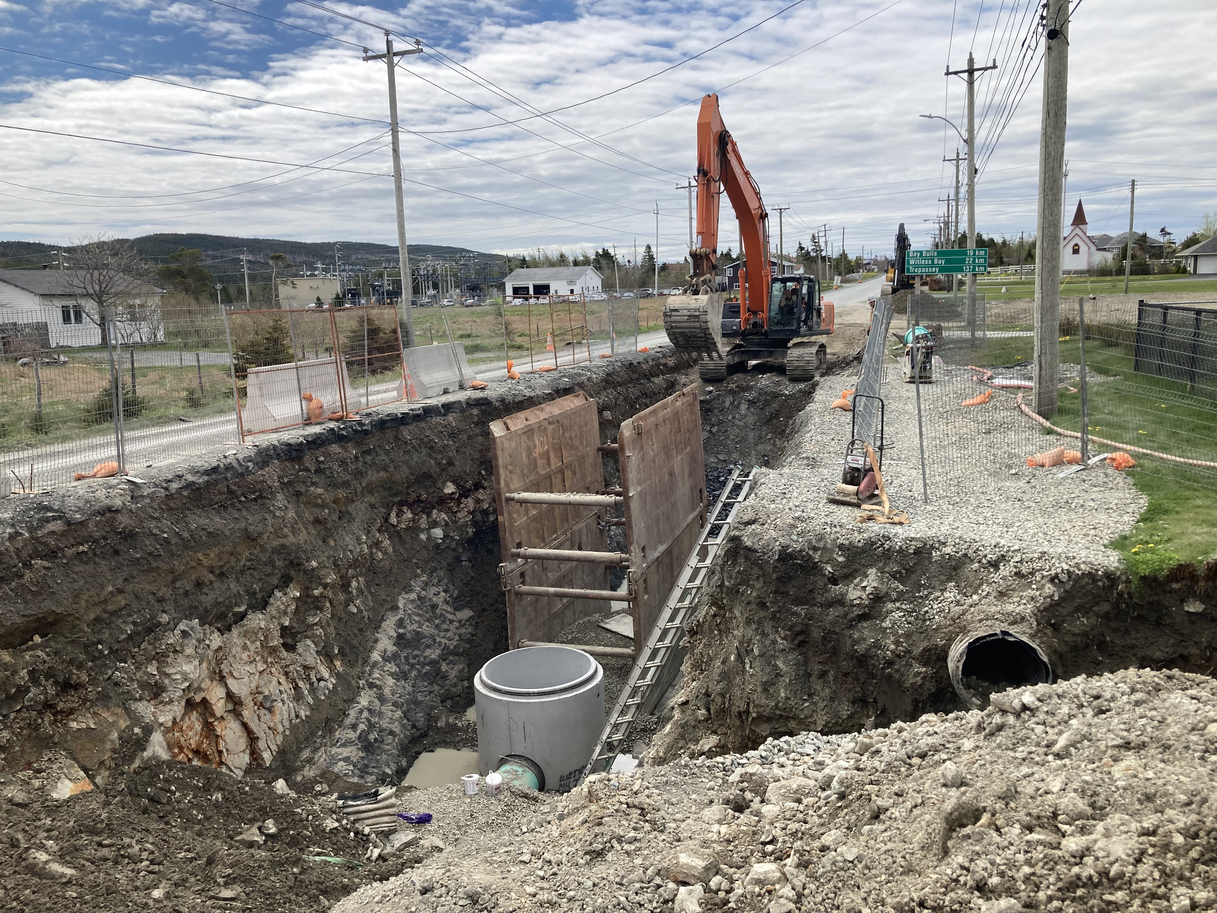 A construction site on a street showing a large hole in the ground, underground piping and a excavator. 