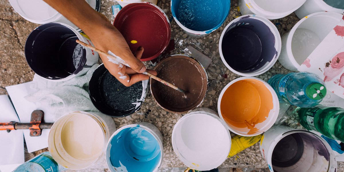persons hand with a paint brush and containers of various colours of paint. 
