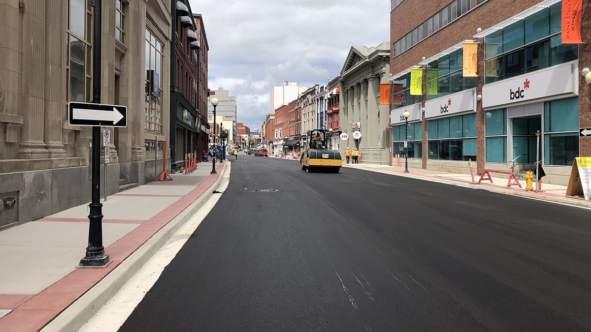 A street scape showing asphalt and construction equipment in the background. There are buildings on both sides of the road.