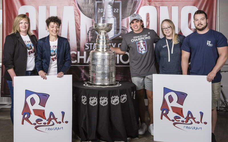 Alex Newhook and REAL Program staff stand on either side of the Stanley Cup.  REAL Program signs in the foreground and a large Colorado Avalanche Stanley Cup Champions creates a backdrop.