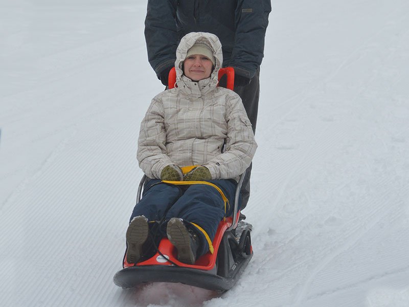 A woman sits in a Snow coach, an adult sized sled that has an upright seat where your legs are extended in front. The snow coach is pushed or pulled by a person other than the rider person