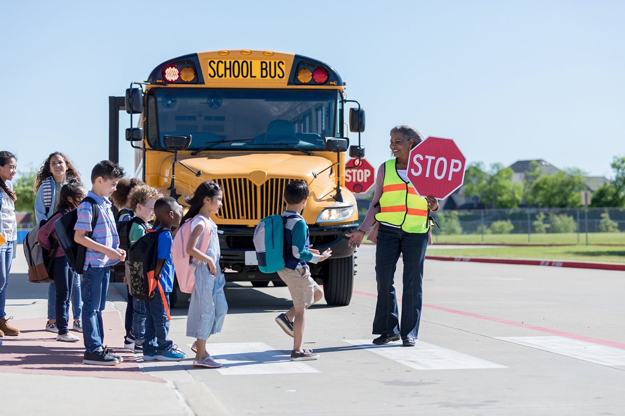 Woman crossing guard holds a stop sign up while children cross on a crosswalk in front of a stopped school bus