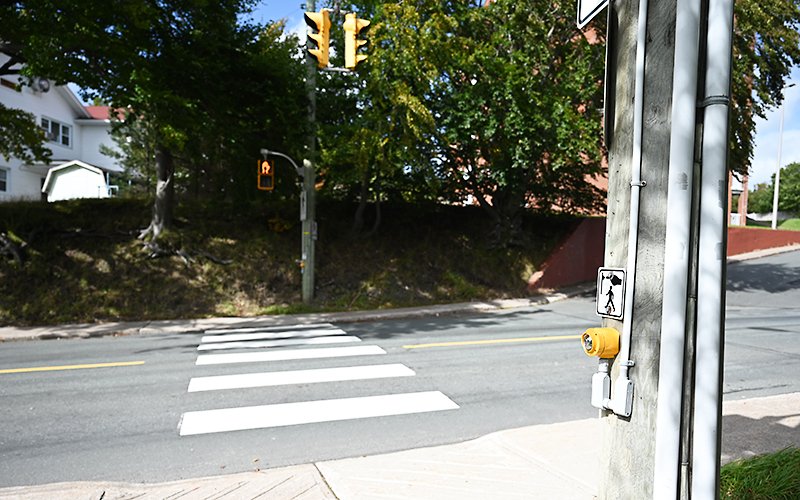 A traffic signal control button mounted on a pole in the foreground with a crosswalk spanning a roadway in the background