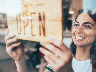 A person smiling while holding a sign saying "Open" through a window. 