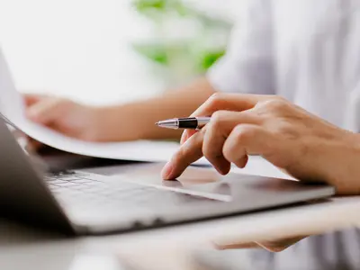 A person using a laptop while holding a sheet of paper.