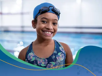 Young girl standing in front of a swimming pool with a swim cap and goggles on.